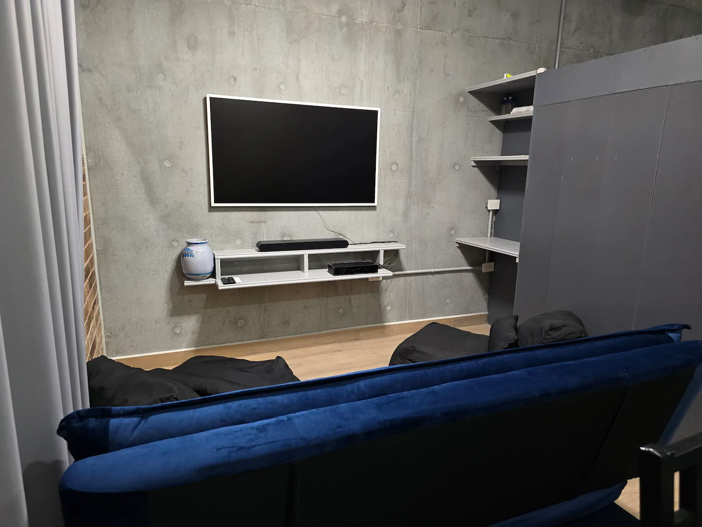 Sala con sofá cama azul, pantalla y muro de concreto